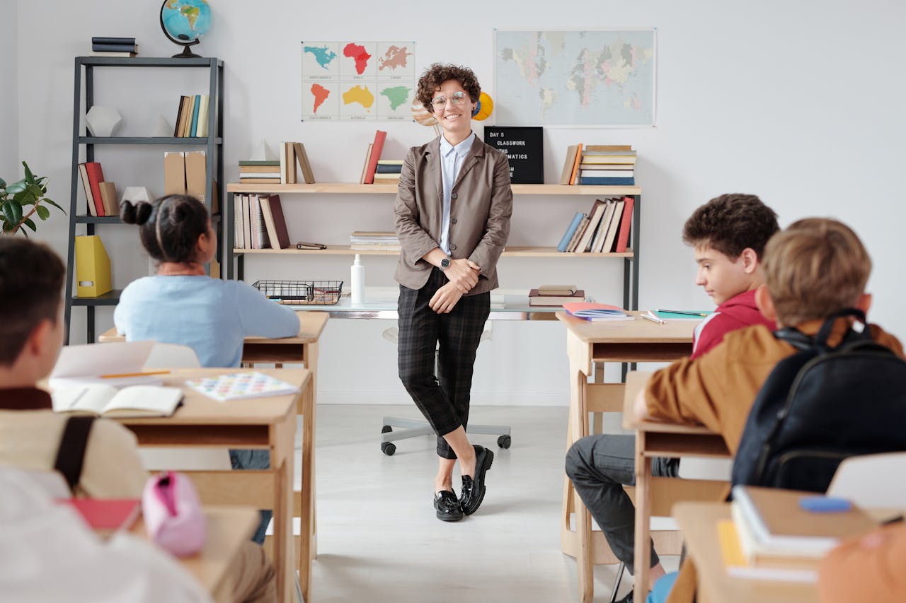 Dans une salle de classe, une enseignante souriante échange avec des élèves assis à leurs bureaux, devant des cartes murales et des étagères de livres.