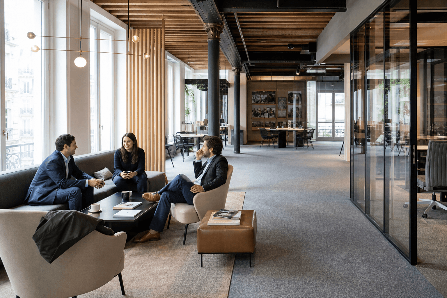 Trois collaborateurs échangent dans un espace de bureaux rénové, avec salon d’accueil, cloisons vitrées et plafond en bois dans un open space.